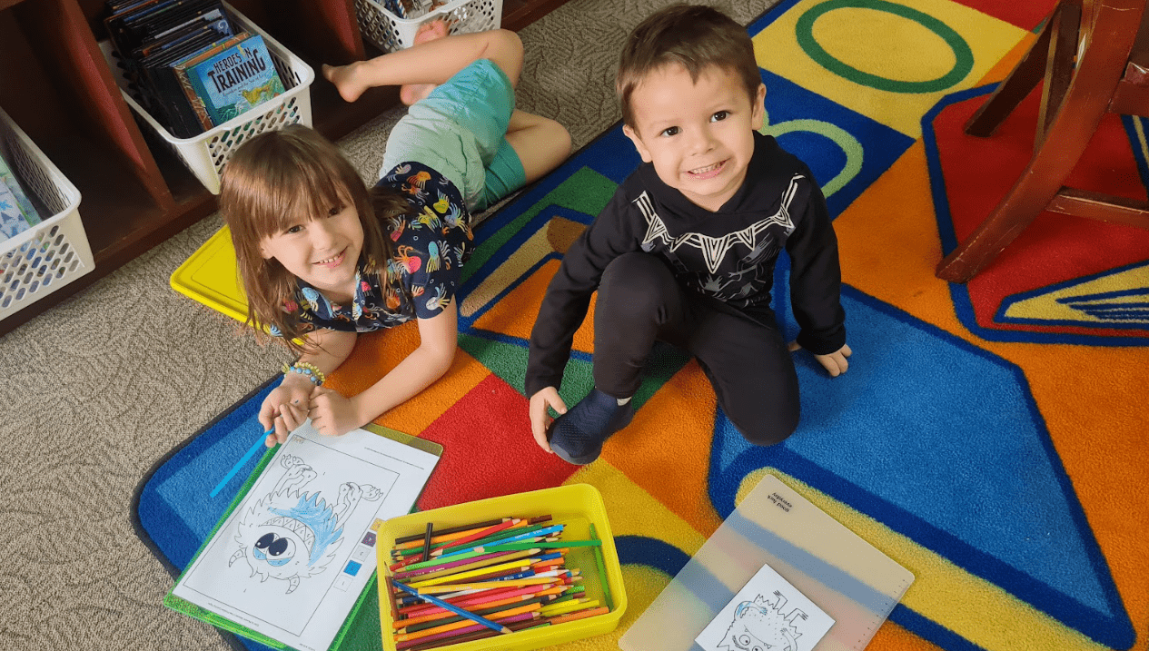 Picture of two kids with coloring monster in a library with books in the background. 